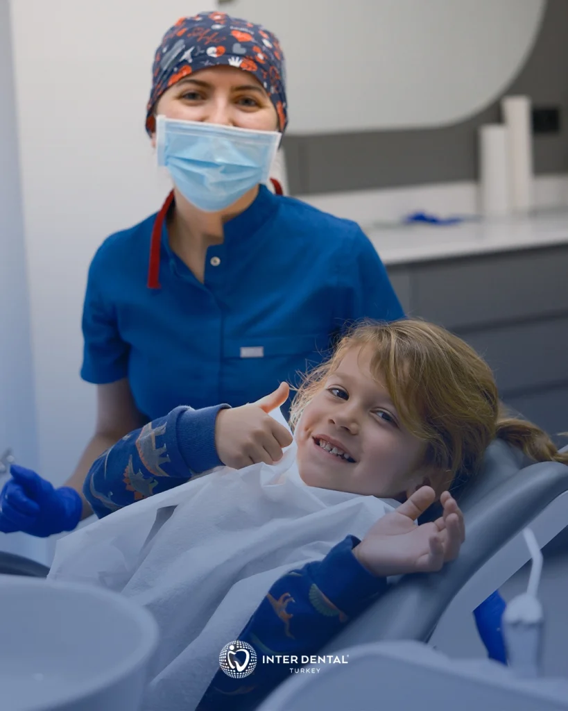 Diş koltuğunda gülümseyen çocuk ve yanında maskeli diş hekimi, çocuk diş hekimliği muayenesi